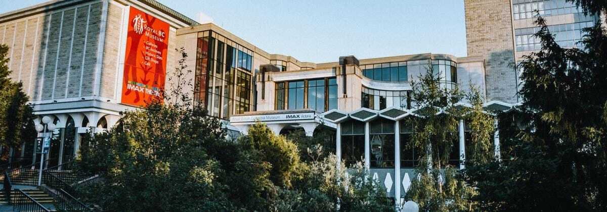 现代建筑配有大玻璃窗，左侧有一面标有「皇家不列颠哥伦比亚省博物馆」的红色横幅和周围的绿色植物。