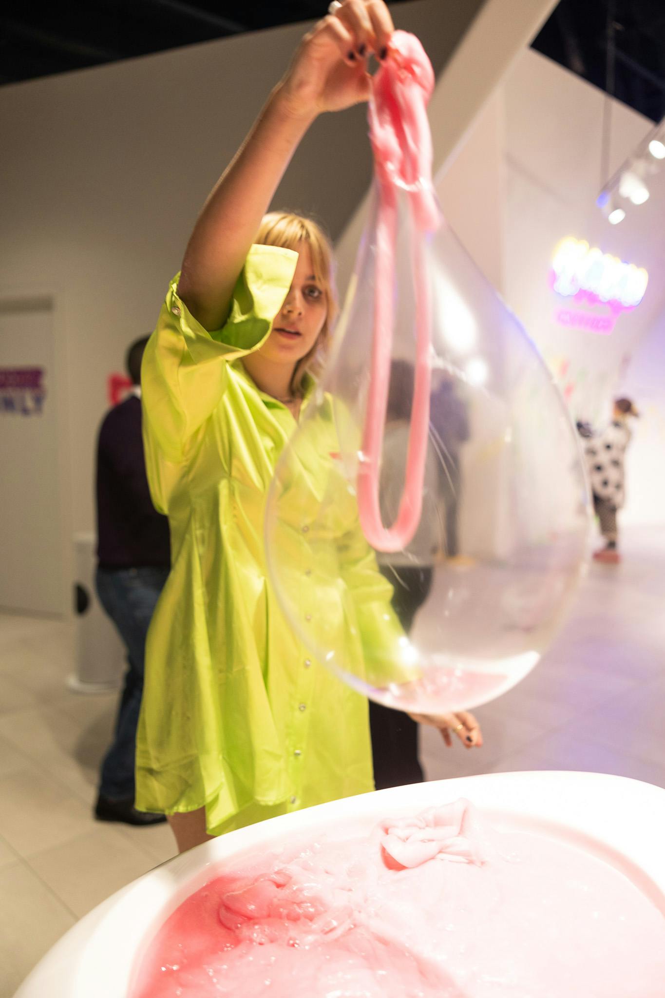 En person i et gult outfit interagerer med en stor, klar ballon i et stærkt oplyst rum med andre mennesker og neonskilte.