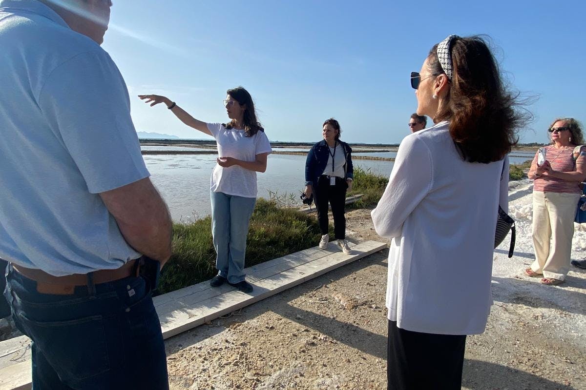 A group of people outdoors near a body of water. One person gestures while others listen and observe the surroundings.