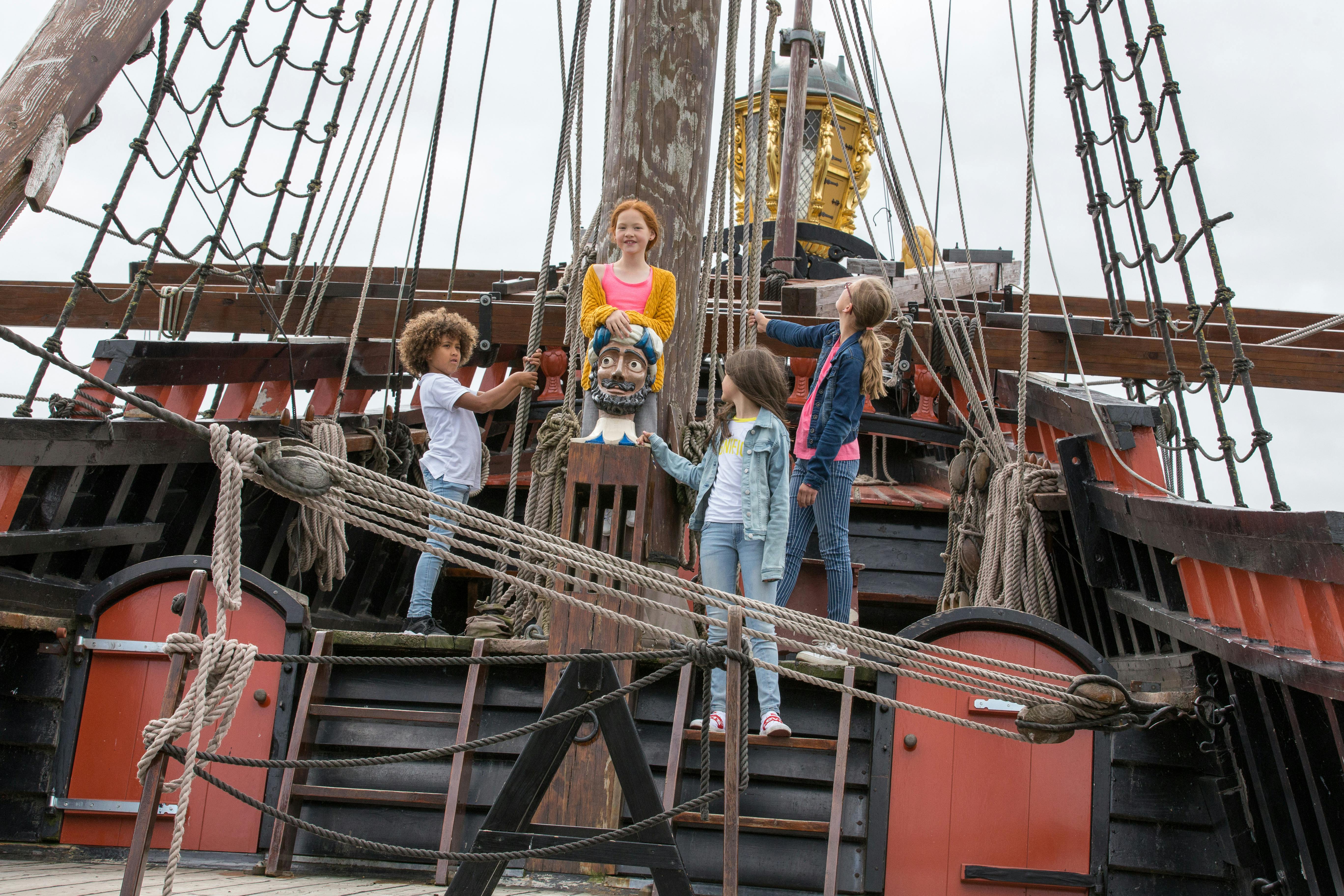 Čtyři děti stojí na palubě staré lodi, obklopené provazy a dřevěnými konstrukcemi.