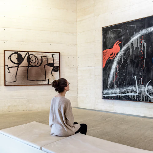 Eine Person sitzt auf einer Bank in einer Galerie und betrachtet zwei abstrakte Gemälde an beigen Wänden.