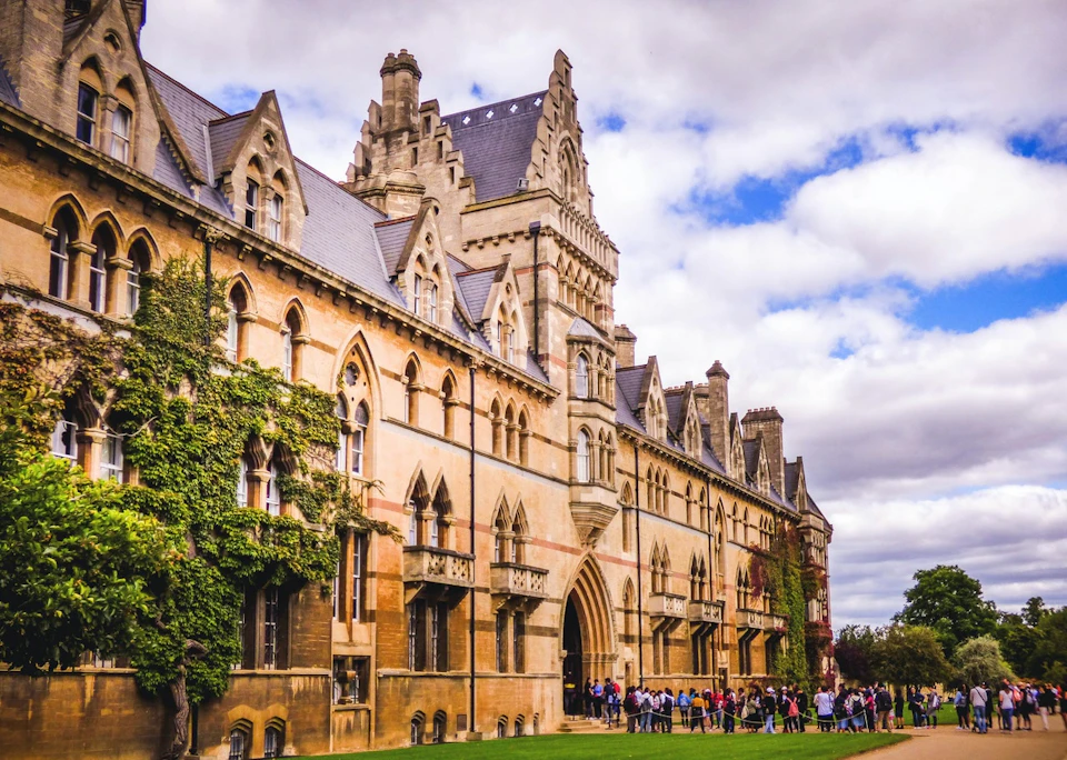 A large, historic building with Gothic architecture. People are gathered near the entrance under a cloudy sky.