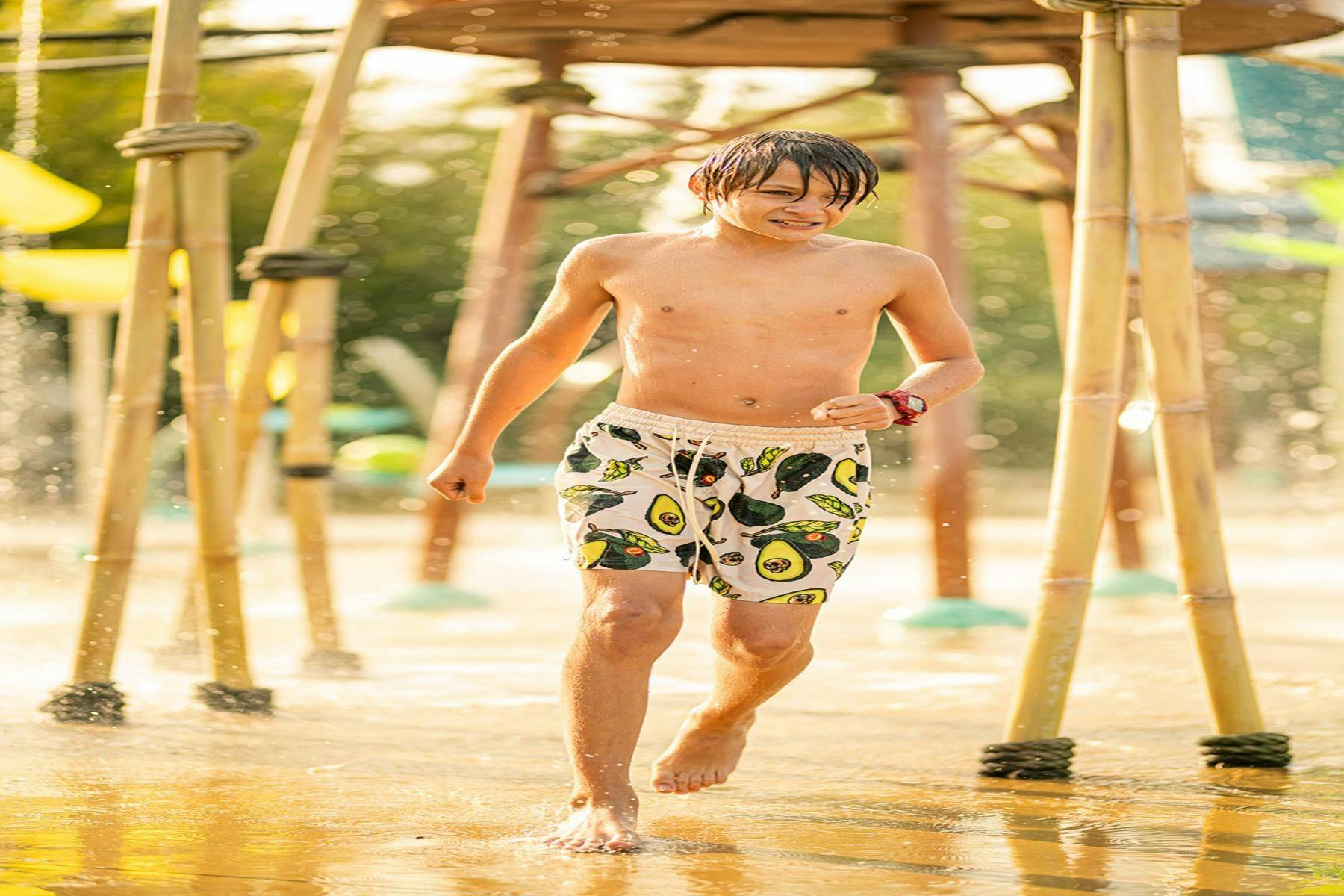 A young person walking through a shallow water area, wearing patterned swim trunks, under a wooden structure on a sunny day.