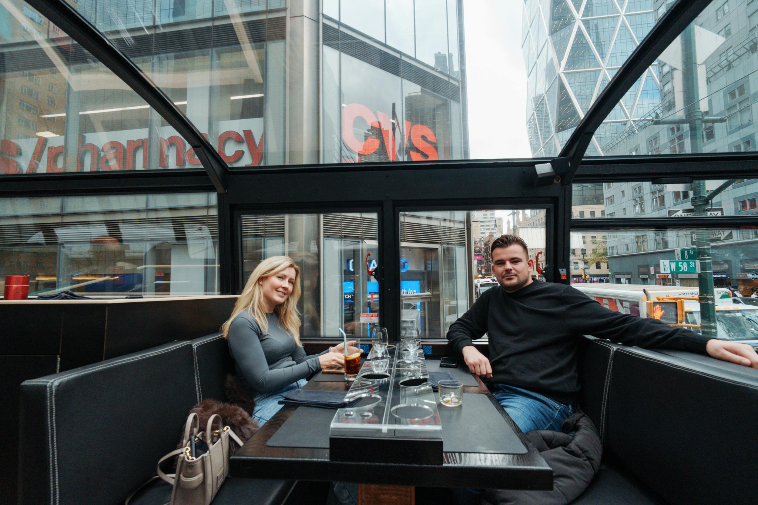 Twee mensen zitten in een glazen restaurant met gebouwen in de stad op de achtergrond, waaronder een CVS-winkel.