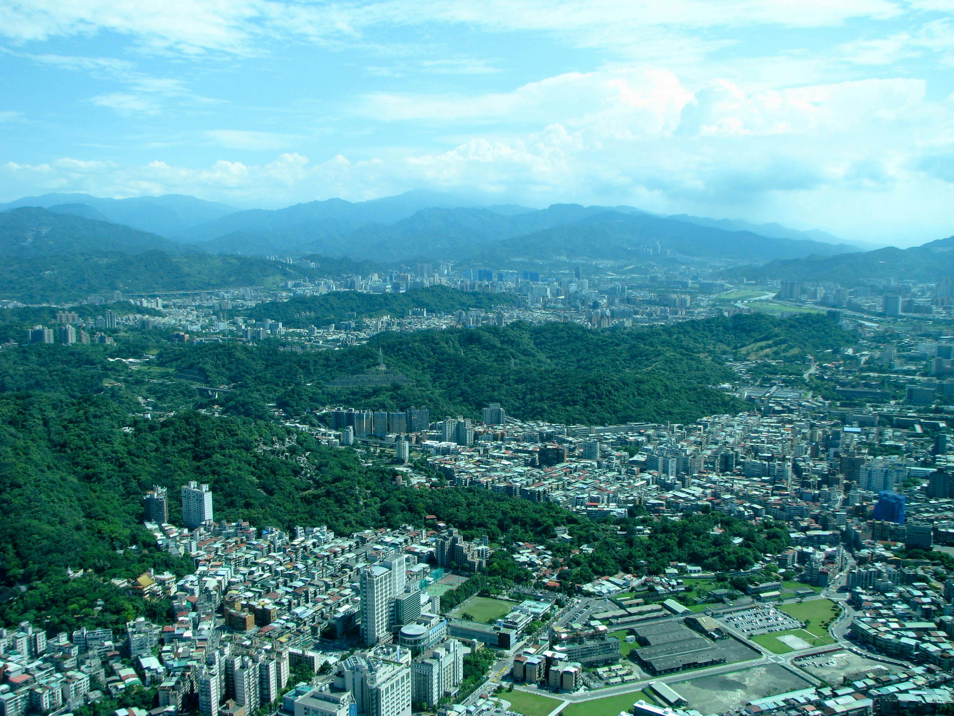 Aerial view of a city with dense buildings, green hills, and mountains in the background under a blue sky with scattered clouds.