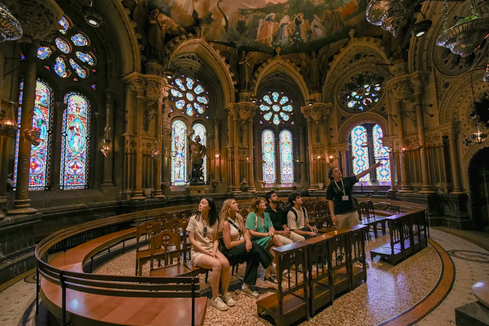 A group of people seated and listening to a guide inside a richly decorated room with stained glass windows and ornate architecture.