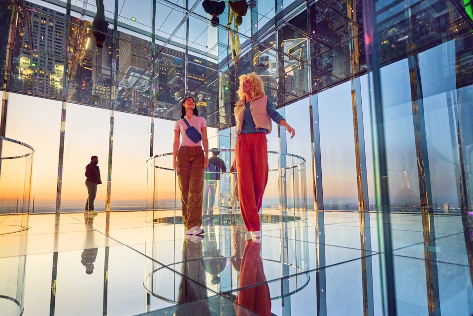 Two people standing in a mirrored observation room with a view of a cityscape during sunset.