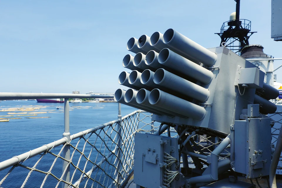 A close-up of a naval ship's missile launcher with multiple cylindrical tubes, overlooking a harbor under a clear blue sky.