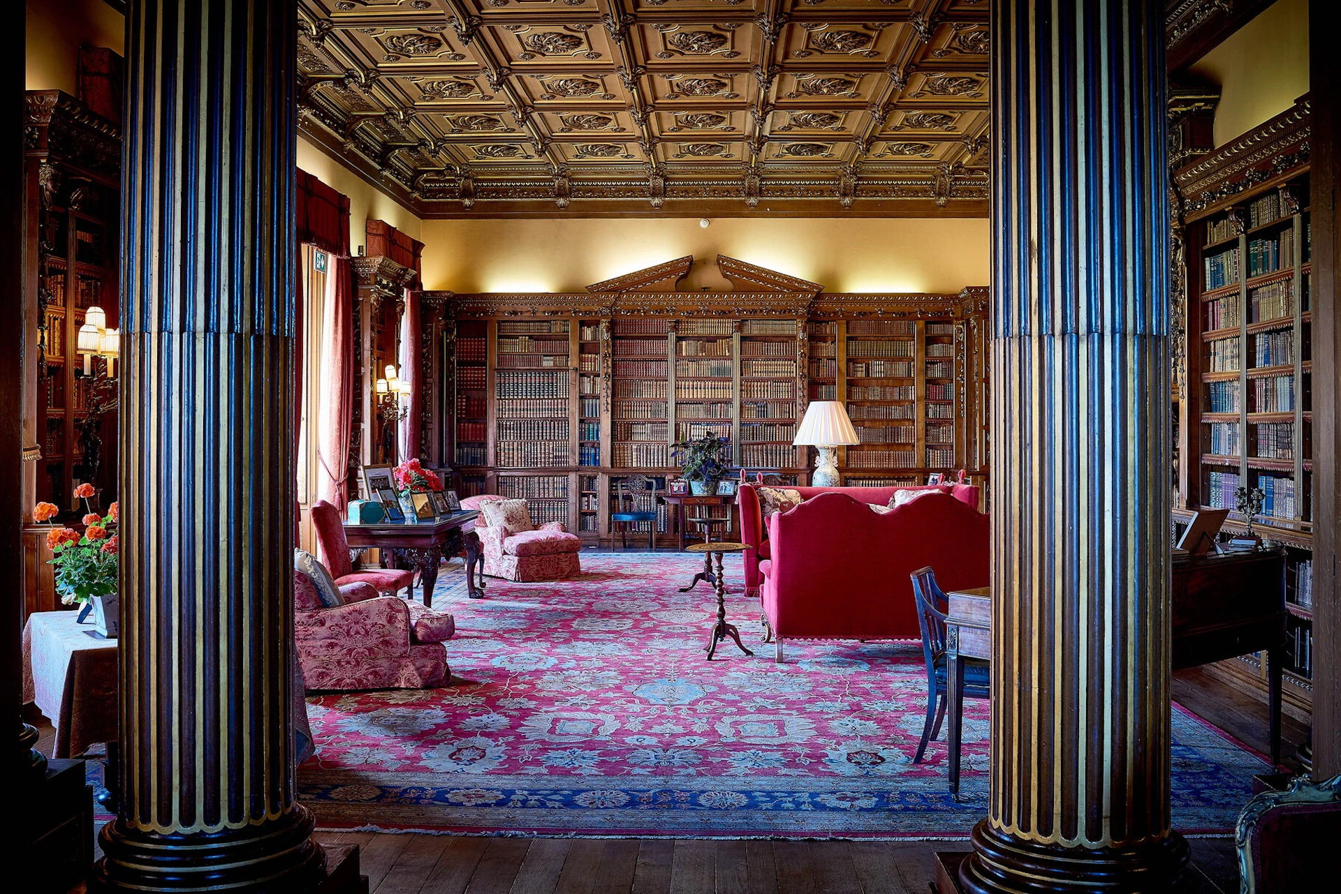 A luxurious library with ornate wood paneling, bookshelves, red upholstered furniture, and a decorative ceiling.