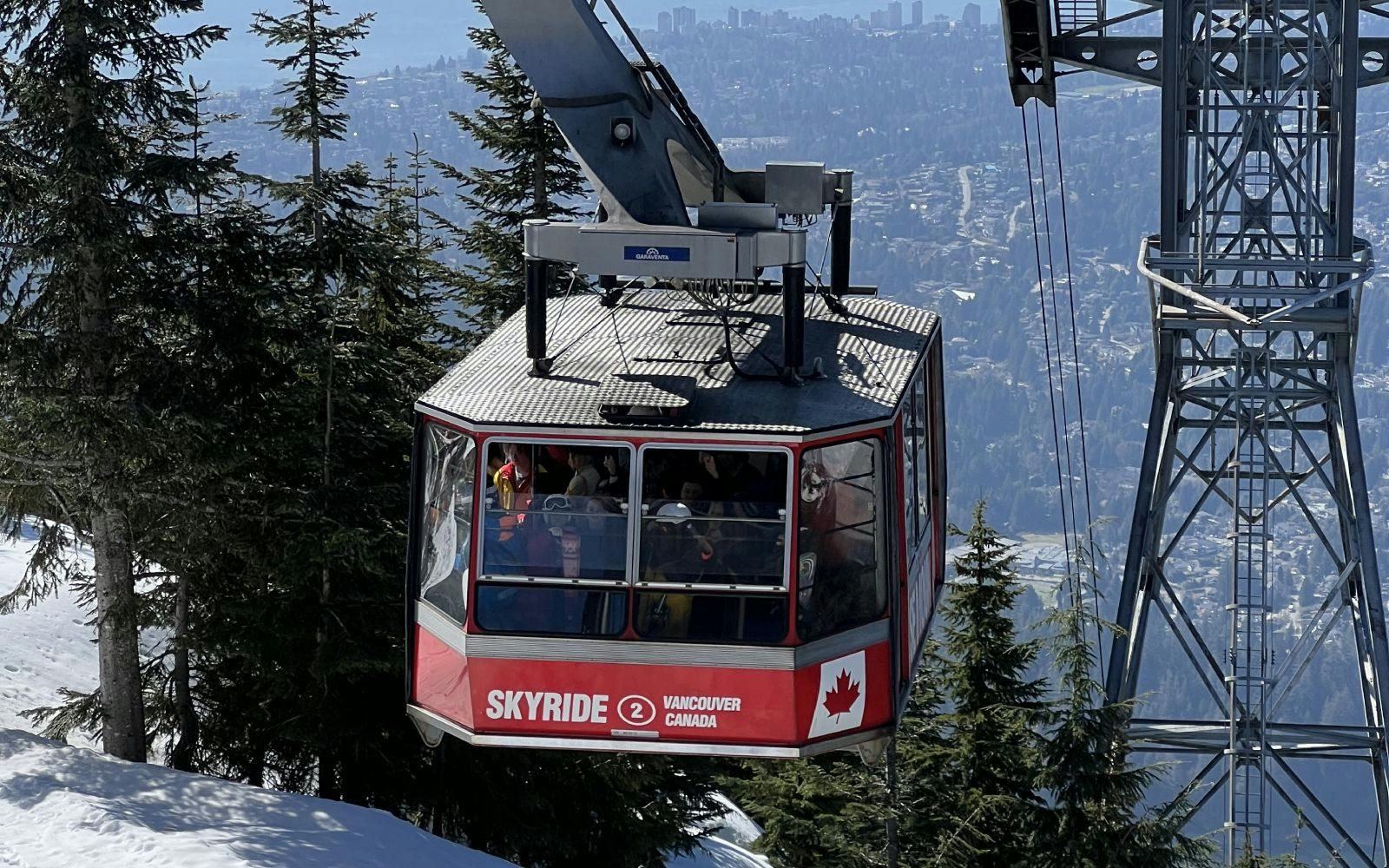 「SKYRIDE」と名付けられた赤と白のゴンドラが、常緑樹が生い茂る雪山を登り、街並みを背景にしています。