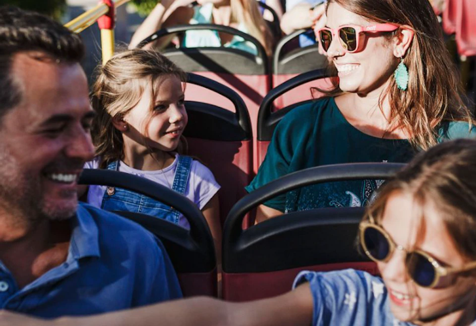 People smiling and enjoying a sunny day on a double-decker bus. A child playfully extends an arm towards an adult.