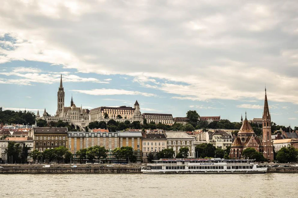 Riverbank cityscape with historic buildings, a prominent church spire, and a docked river cruise ship under a cloudy sky.