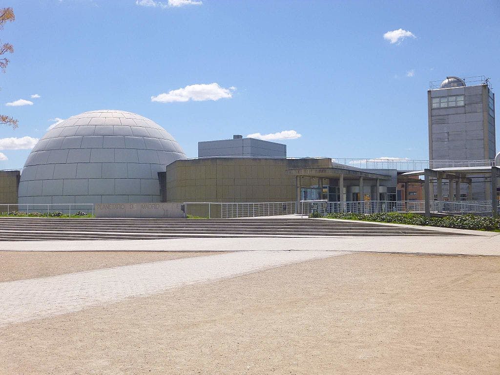 Edificio moderno con una gran estructura en forma de cúpula, plaza abierta con escalones y cielo azul despejado. El texto del edificio dice "Planetario de Madrid".