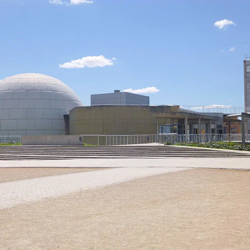 Modern byggnad med en stor, kupolformad struktur, öppet torg med trappor och klarblå himmel. Texten på byggnaden lyder "Planetario de Madrid".
