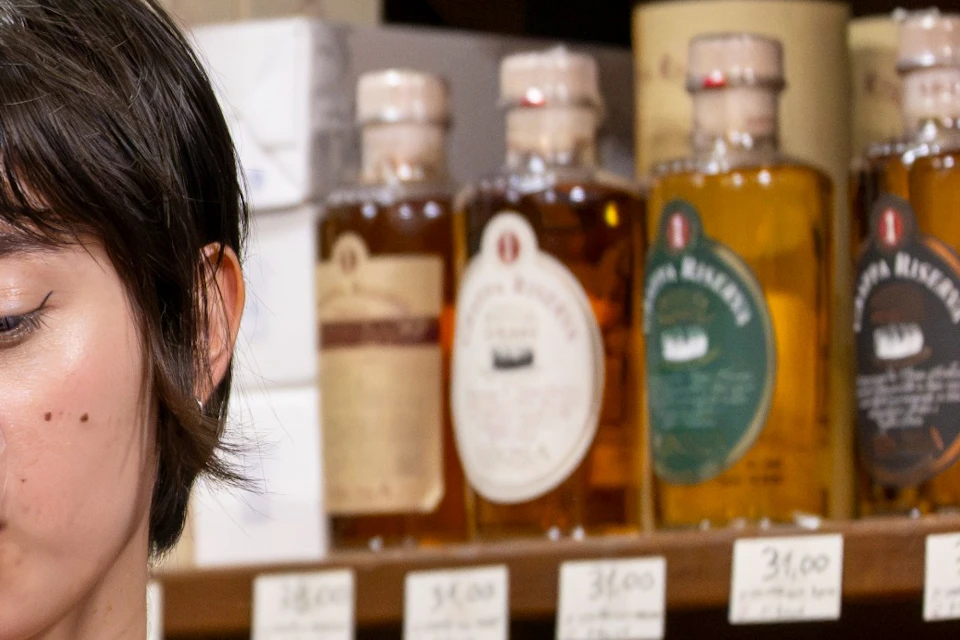 A person in a red and white top tasting red wine in a store with shelves of assorted bottles in the background.
