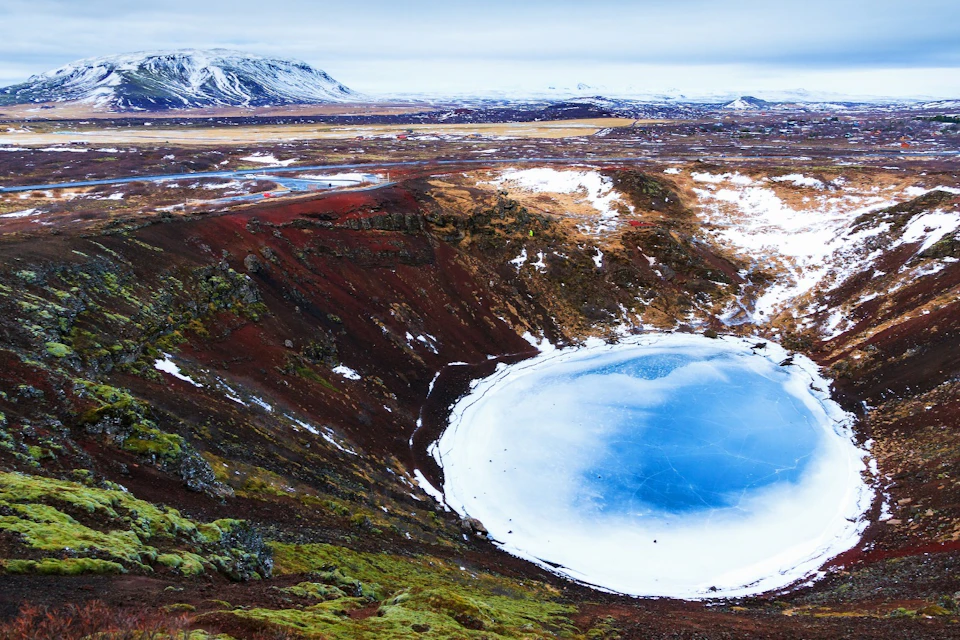 Kerið Crater in winter with a mountain in the background. Snow, green moss, red rocks.