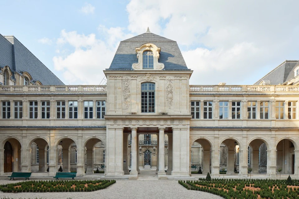 A neoclassical building with arched windows and elaborate stonework, featuring a central doorway and manicured garden in front.