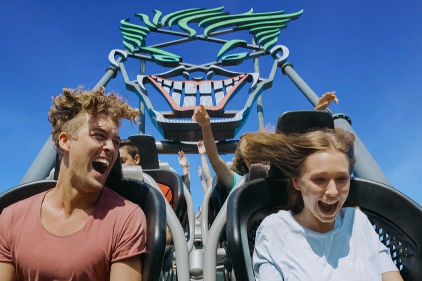 Two people scream with joy on a rollercoaster with a grinning face design in the background under a clear blue sky.