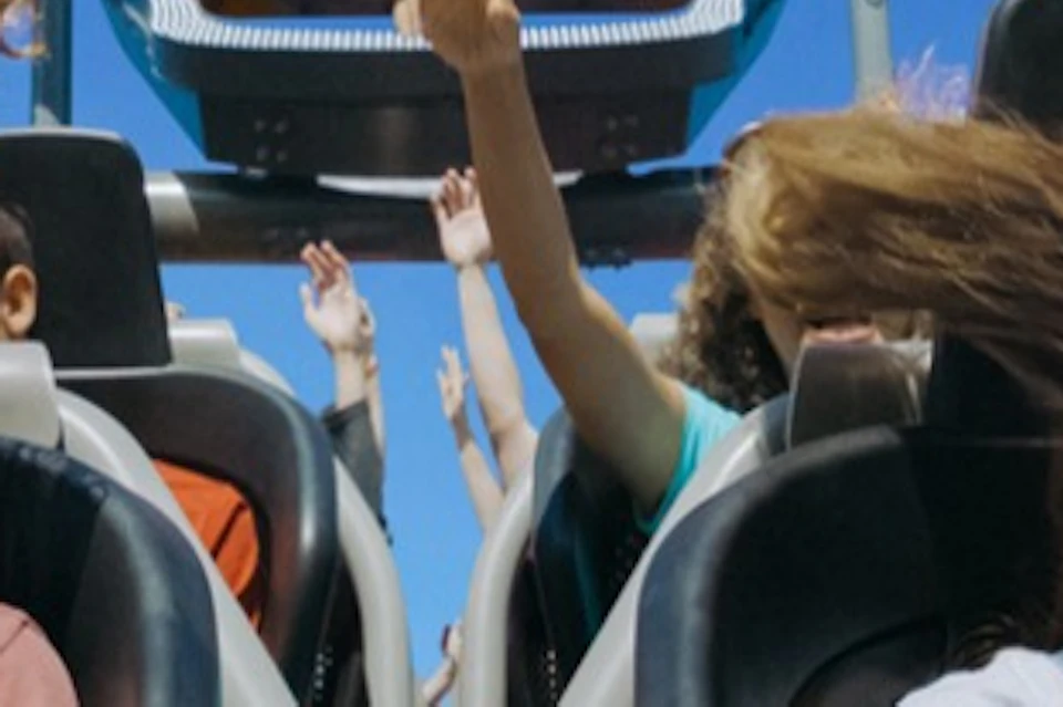 Two people scream with joy on a rollercoaster with a grinning face design in the background under a clear blue sky.