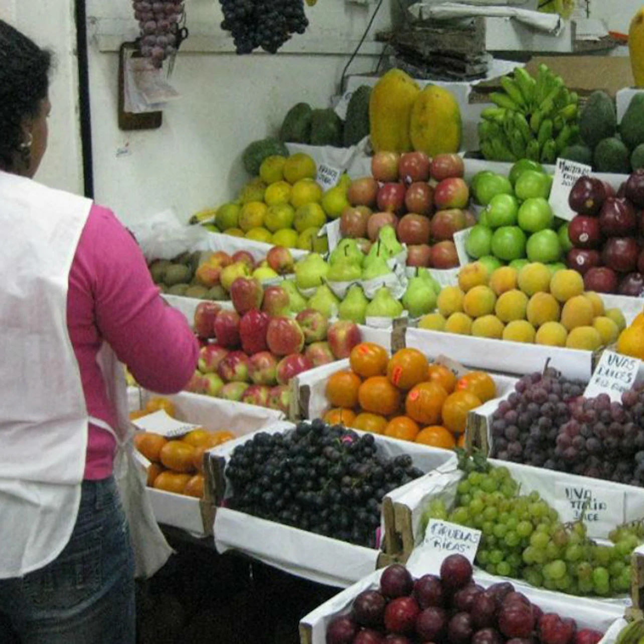 A person in a pink shirt and white apron arranging various fruits, such as apples, oranges, and grapes, at a market stall.