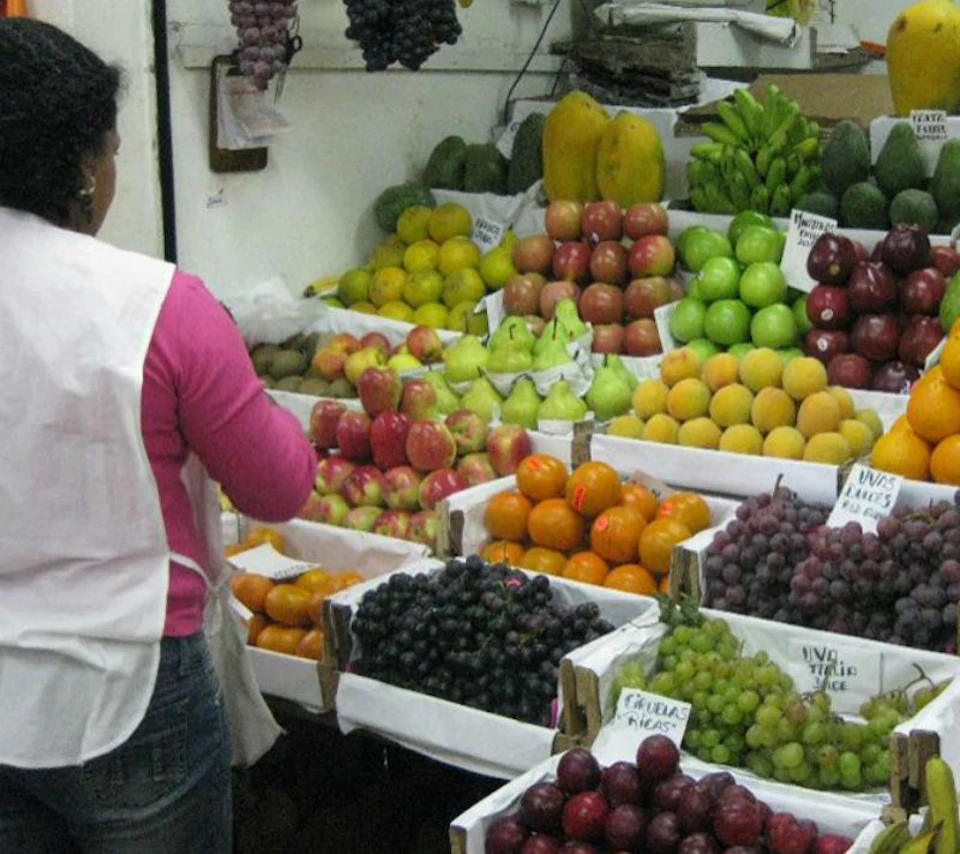 A person in a pink shirt and white apron arranging various fruits, such as apples, oranges, and grapes, at a market stall.