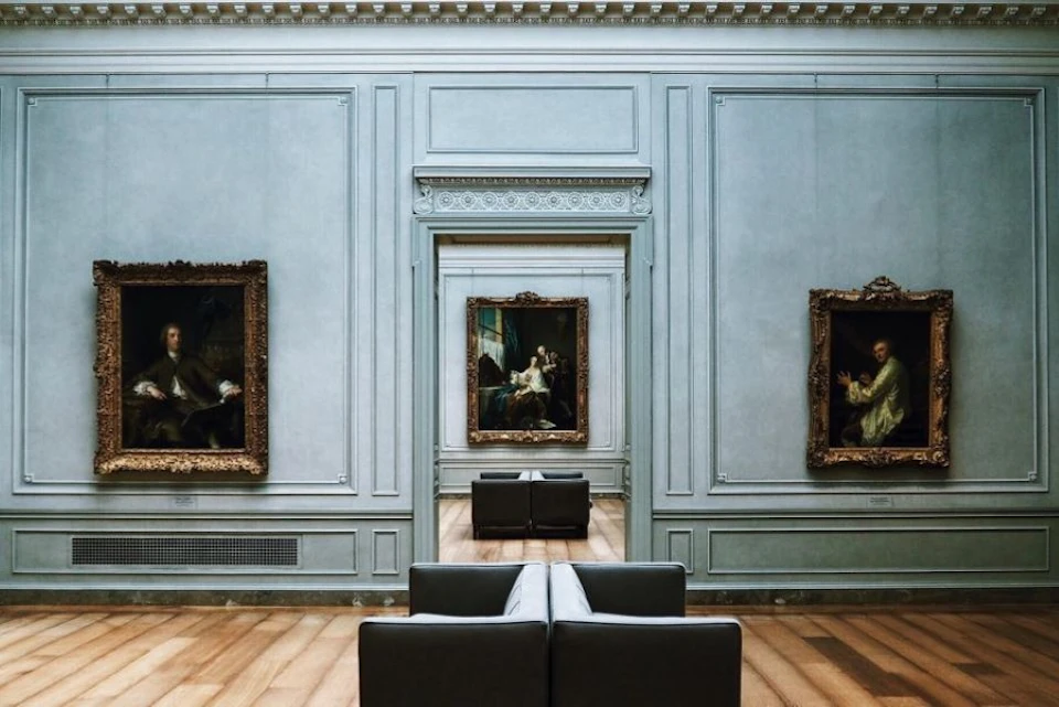 Art gallery room with ornate framed paintings on light blue walls, empty gray seating arrangement on wooden floor in the center.