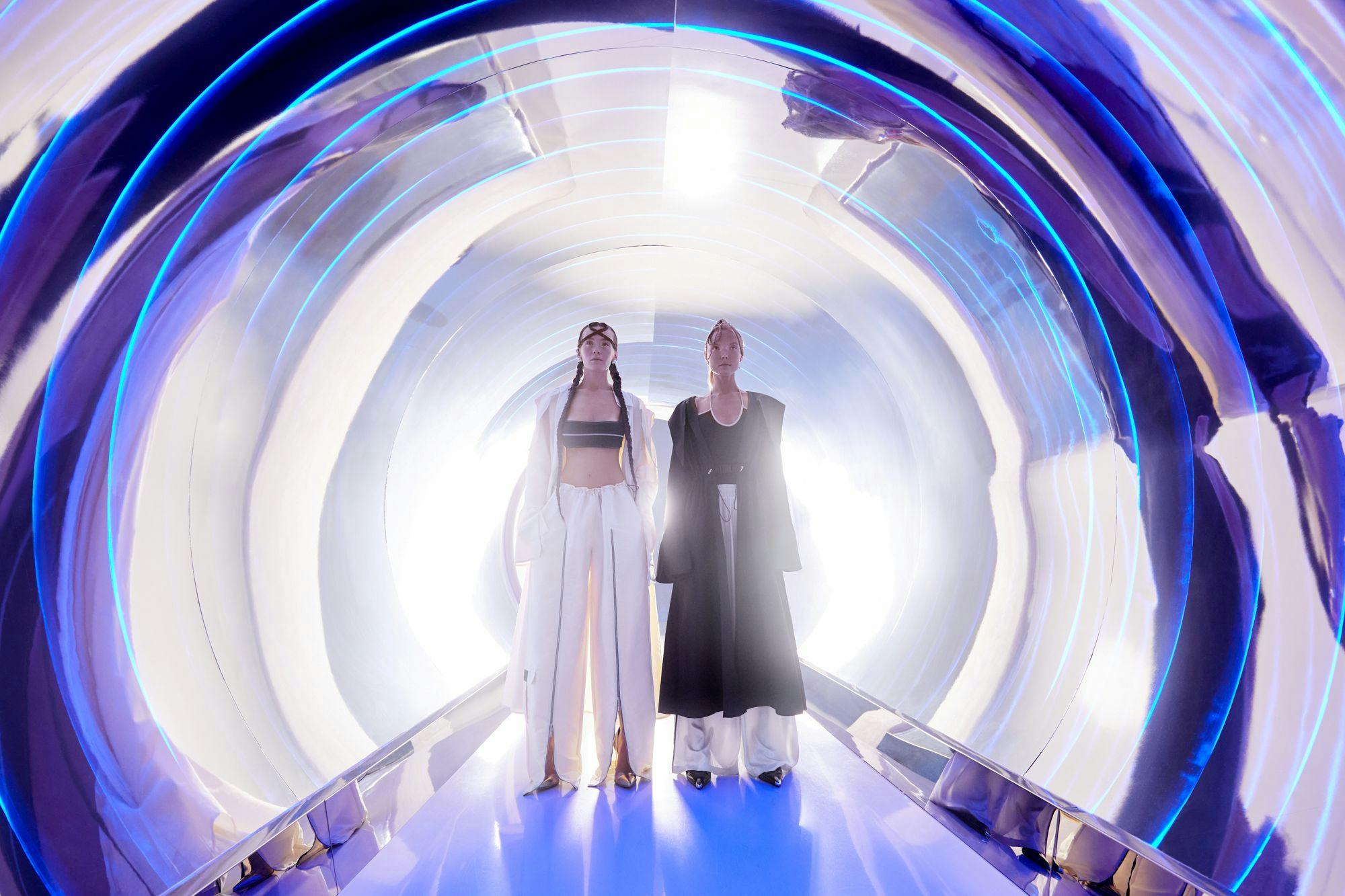 Δύο άνθρωποι με φουτουριστικά ρούχα στέκονται σε ένα λαμπερό τούνελ με μπλε και λευκά φώτα.