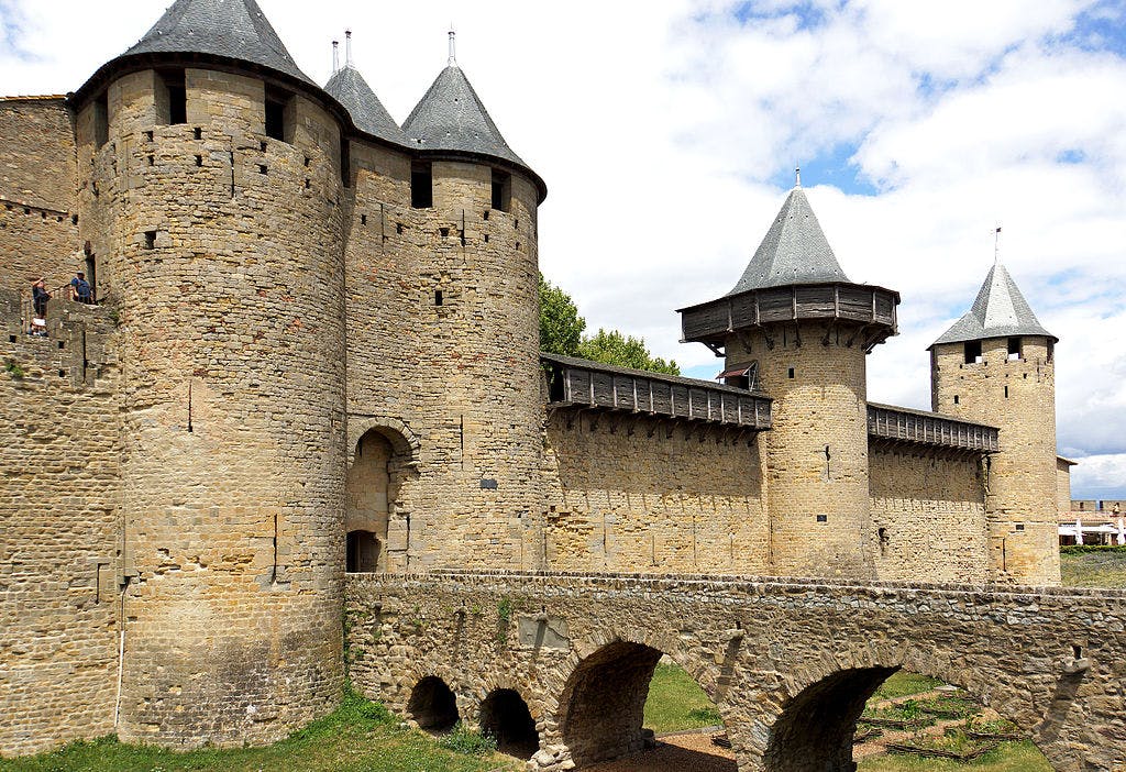 Medieval stone castle with four round towers, a defensive wall, and an arched bridge. Two people are standing on the left.