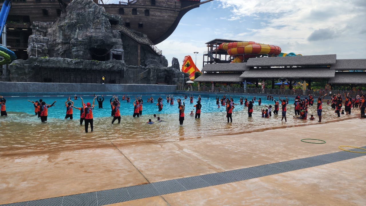 身着红色和黑色制服的人们正在波浪池中引导一群人，背景是一艘海盗船和五颜六色的水滑梯。