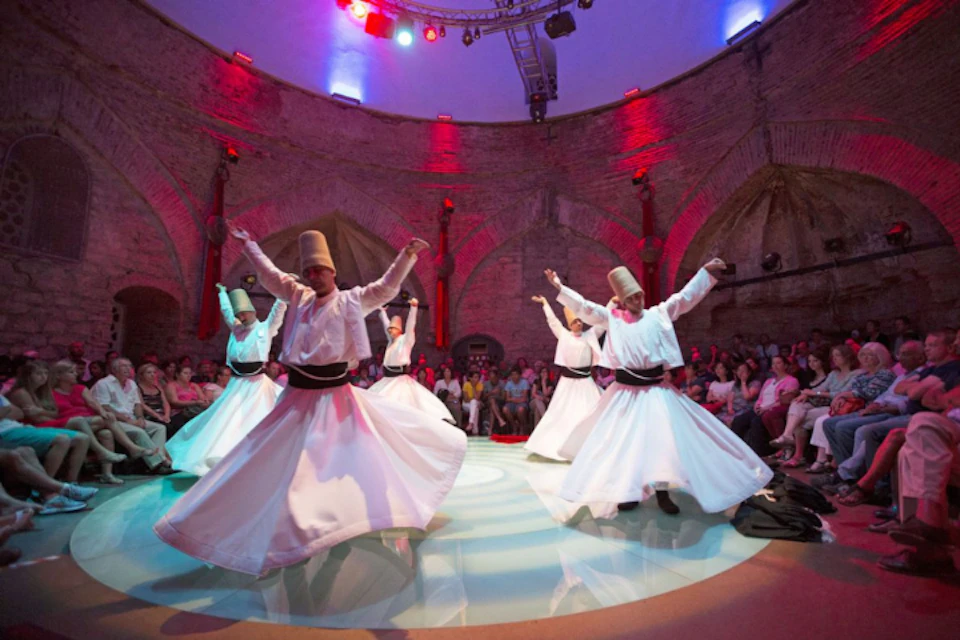 Dancers in white robes and conical hats performing in a circular, illuminated room with an audience seated around them.