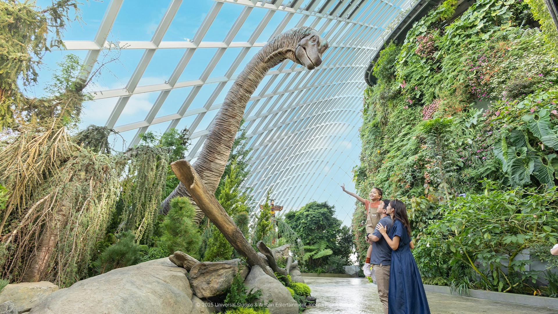 한 가족이 유리 천장이 있는 울창한 실내 식물 환경에서 키가 큰 공룡 모델을 관찰하고 있습니다.