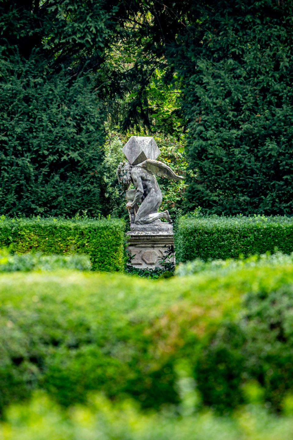 A stone statue of a winged figure kneeling with a box on its back, surrounded by dense green hedges and trees.