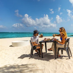 Two people sitting at a small table on a sandy beach, enjoying drinks and food under a clear blue sky with ocean in the background.