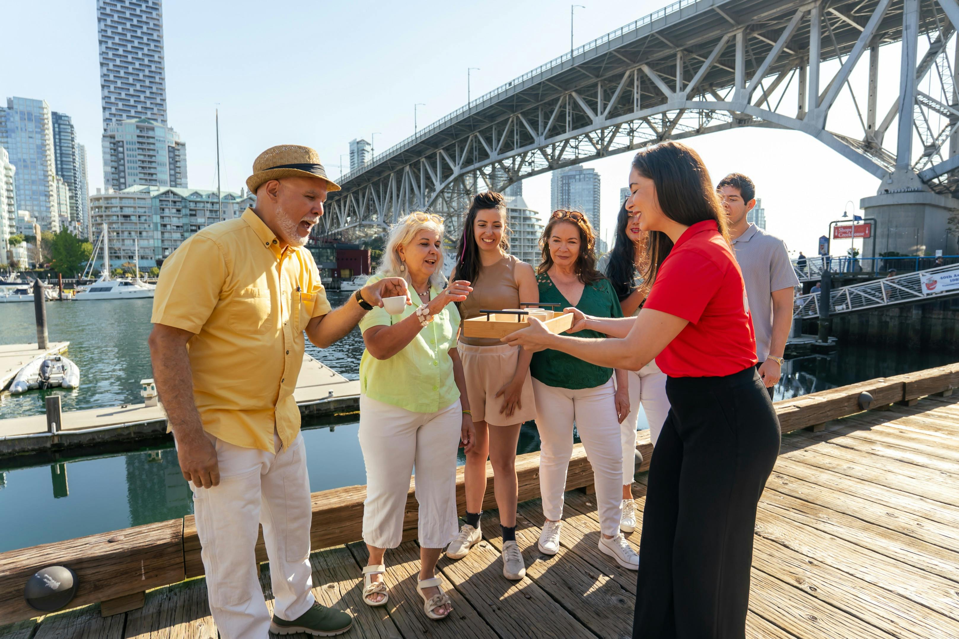 커다란 다리 아래 선착장에 있는 한 무리의 사람들이 빨간 셔츠를 입은 여성으로부터 작은 접시 쟁반을 받고 있습니다.