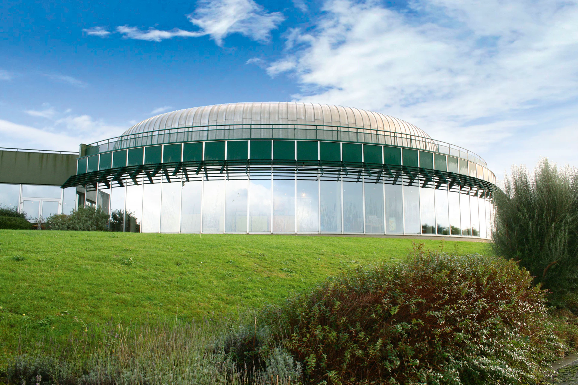 A dome-shaped, glass-walled building with a green base, set against a cloudy blue sky, surrounded by green grass and shrubs.