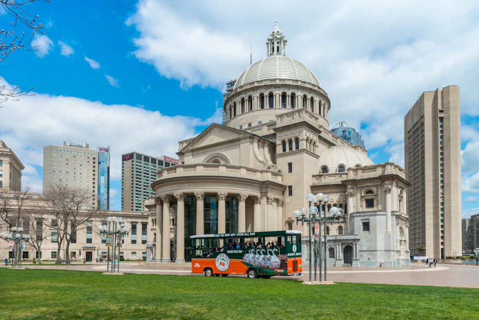 A large, domed classical building under a partly cloudy sky with a passing green and orange tour bus in the foreground.