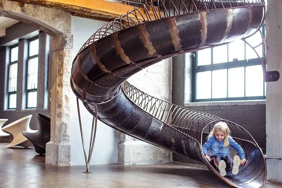 Ένα μικρό παιδί με μπλε μπουφάν γλιστράει κάτω από μια εσωτερική σπειροειδή διαφάνεια σε ένα δωμάτιο βιομηχανικού στιλ με μεγάλα παράθυρα.