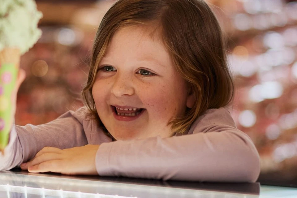 Two smiling children hold ice cream cones while standing at a counter inside a brightly lit shop.