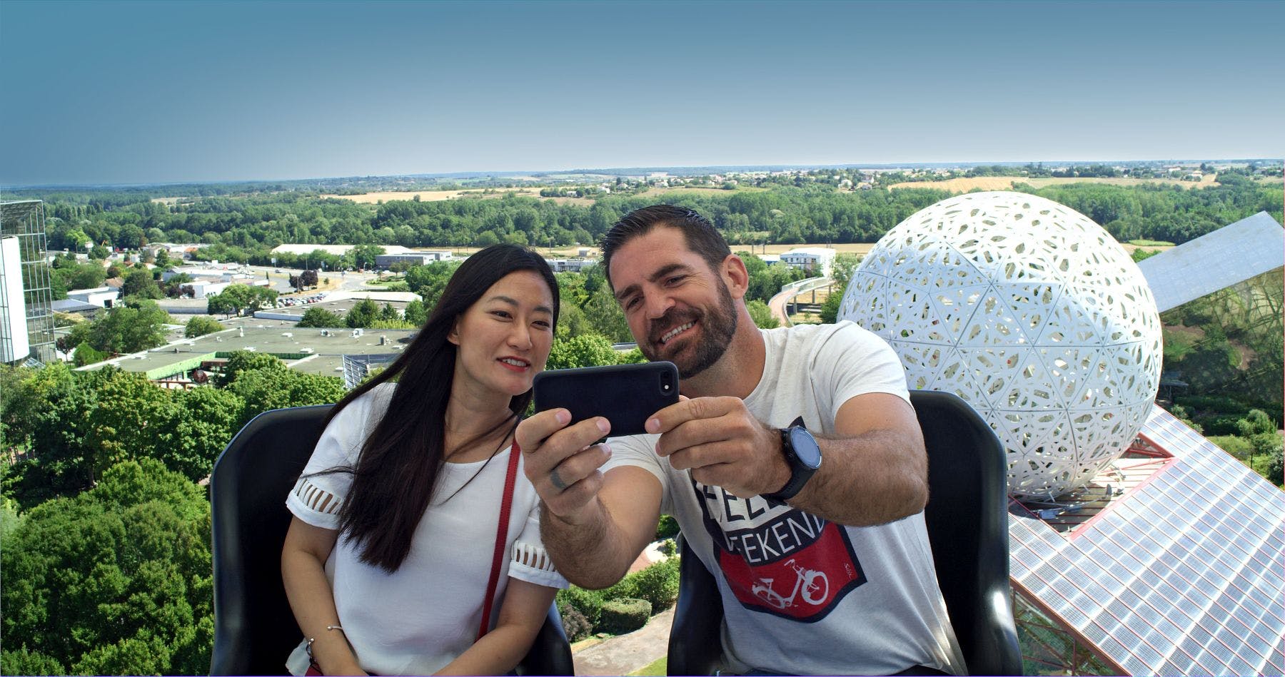 A man and a woman take a selfie while seated above a landscape with a mix of greenery, buildings, and a white dome.