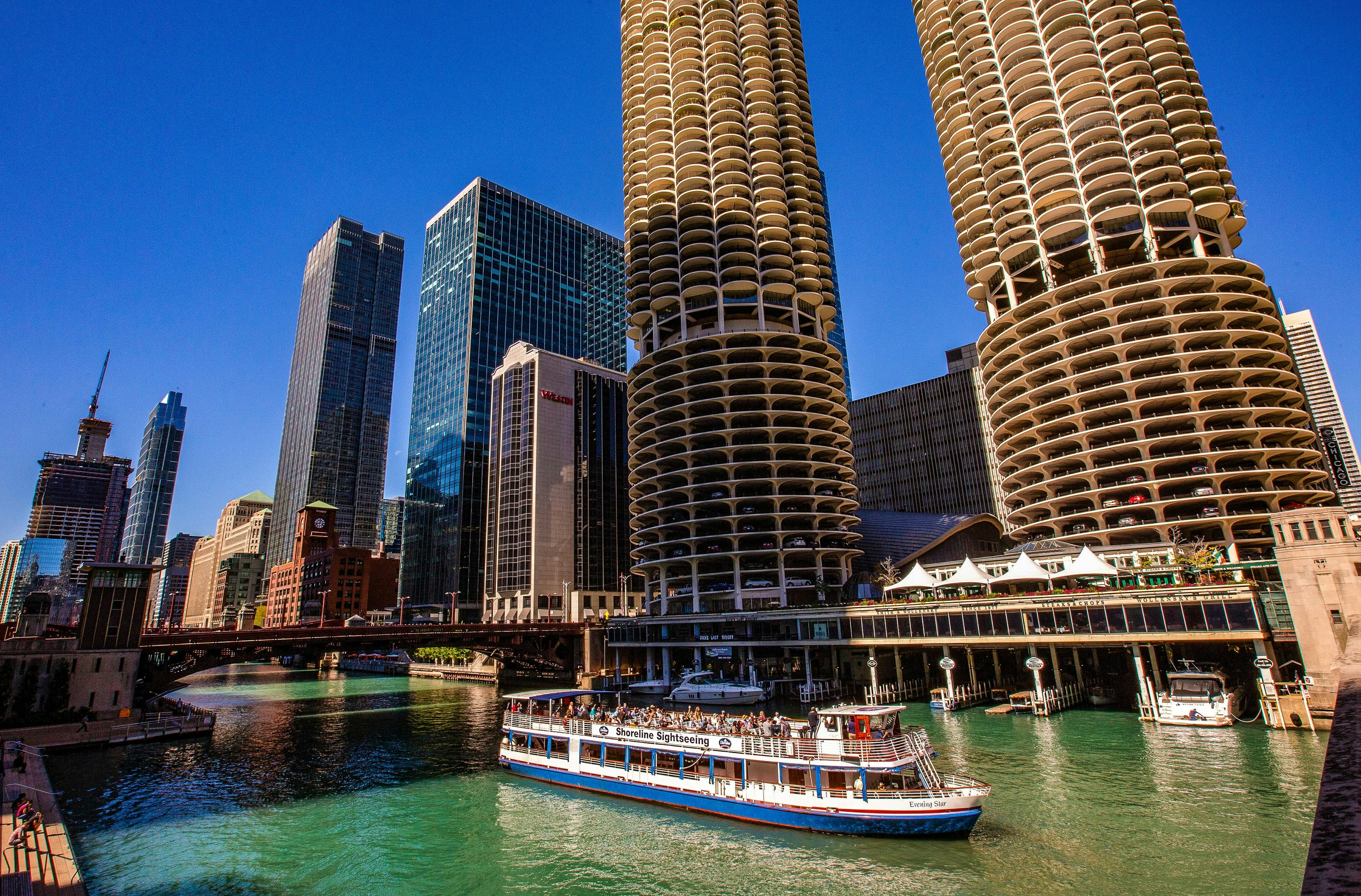 A tour boat cruises on a river in an urban area, surrounded by tall, uniquely cylindrical buildings and skyscrapers.