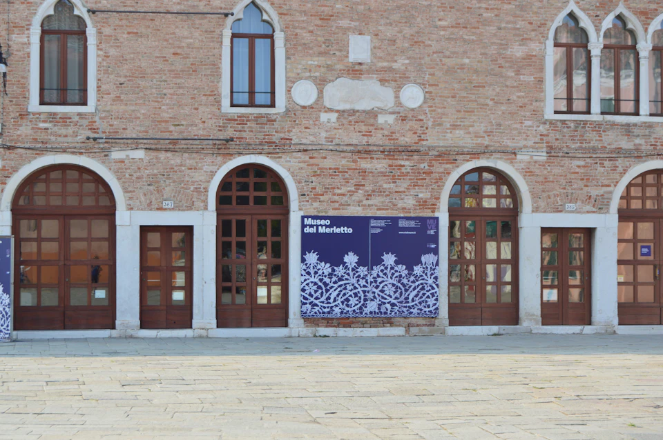 Facade of the Museo del Merletto, showcasing arched windows and a large purple banner with white lace designs.