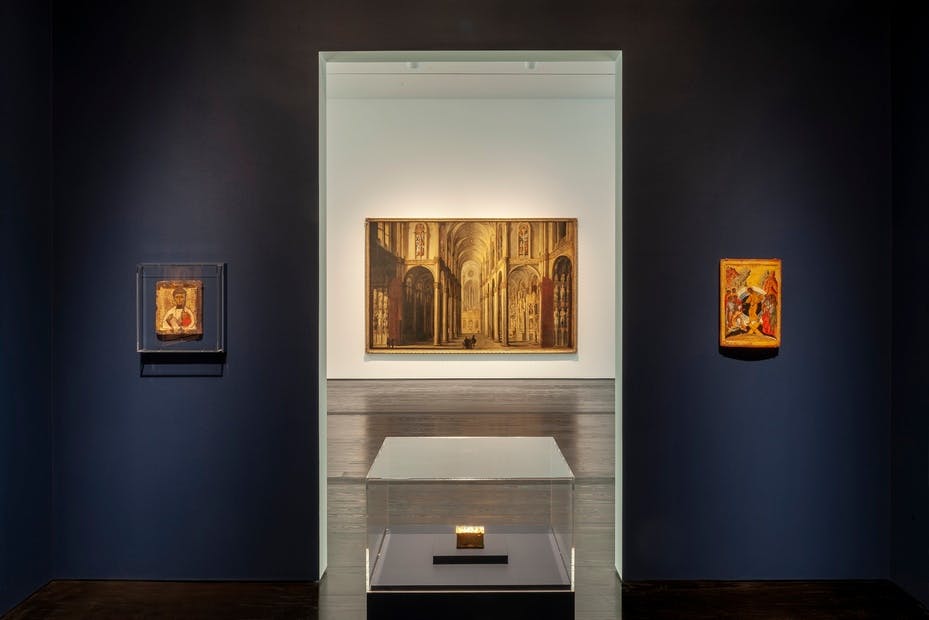 유리 케이스에 담긴 조명 필사본과 짙은 파란색 벽에 그려진 그림 등 종교 예술 작품이 전시된 박물관 갤러리입니다.
