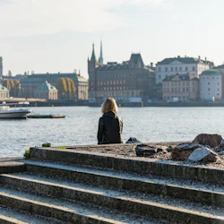 En person sitter på stentrappor vid vattnet, vänd mot en stadsbild med historiska byggnader och en båt på floden.
