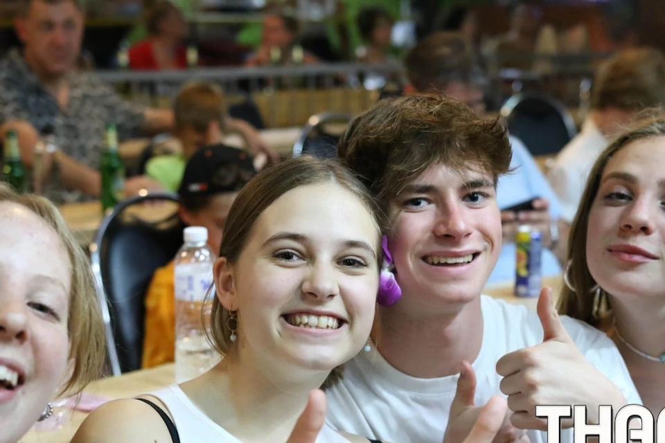 A group of young people smiling and giving thumbs up at a casual indoor event, with others seated in the background.