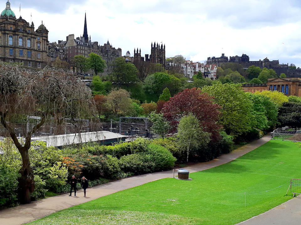 A park with pathways and lush trees, overlooked by historic buildings and church towers under a cloudy sky. Two people walk on a path.