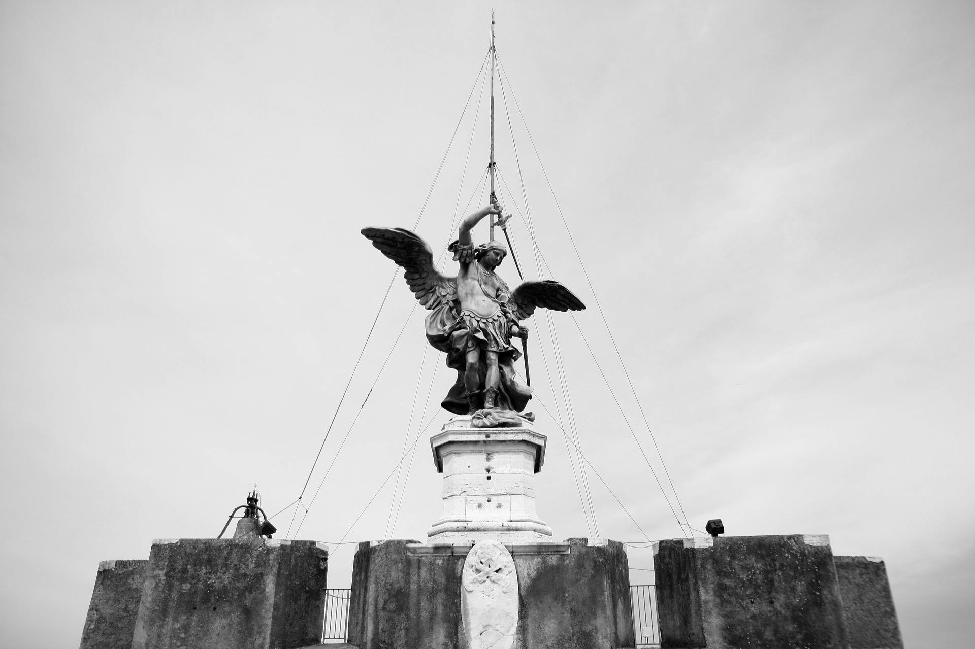 Statue einer geflügelten Figur, die ein Schwert hält, auf einem Sockel, umgeben von einer Steinbalustrade und Kabeln, in Schwarz-Weiß.