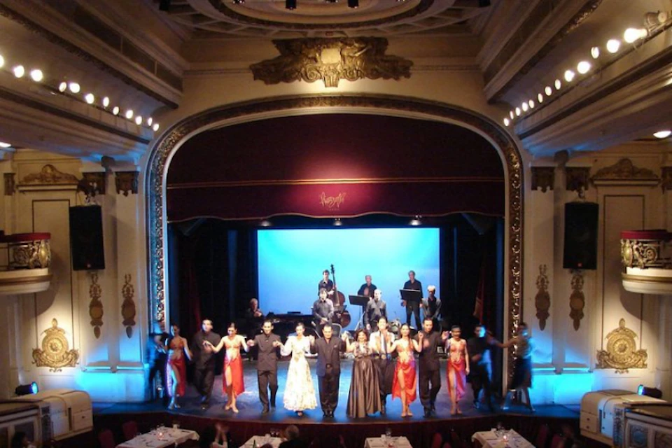 Performers and musicians on a large, ornate stage take a bow in front of a blue backdrop in an elegant theater.