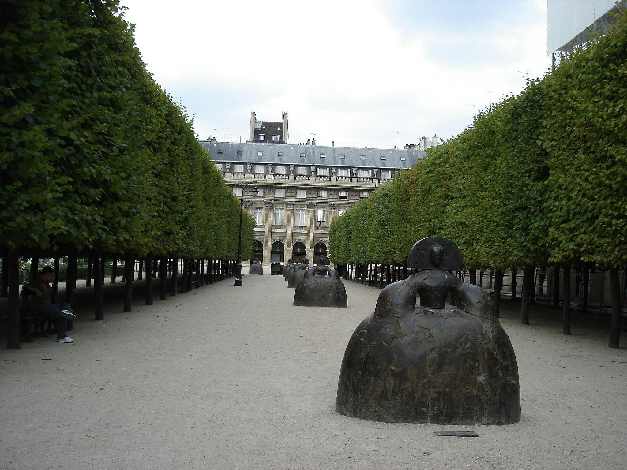 Caminho arborizado com uma série de esculturas abstractas de grandes dimensões, que conduz a um grande edifício. Uma pessoa senta-se num banco à esquerda.