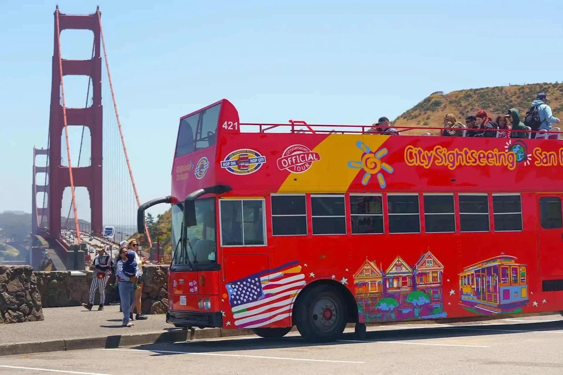 Red double-decker tour bus with passengers, parked near the Golden Gate Bridge on a sunny day.