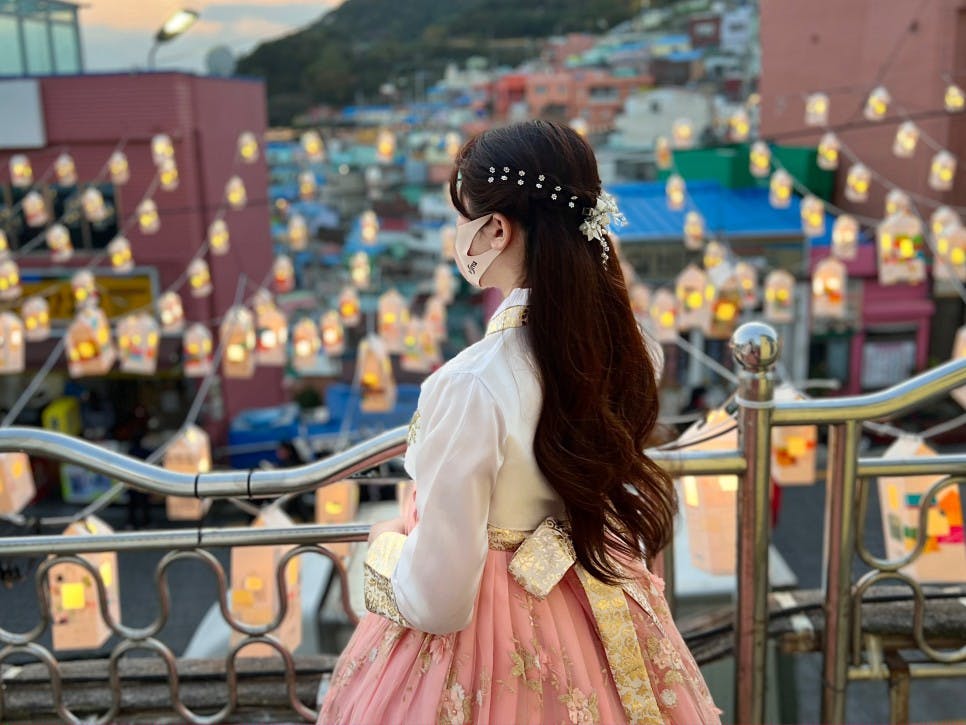 Uma mulher, vestida com um traje tradicional e uma máscara, está numa varanda com vista para uma cidade decorada com lanternas suspensas.